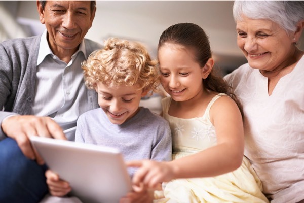 Happy multi-generational family — grandparents and two young grandchildren — gathered closely on a couch, smiling while looking at a tablet screen together.