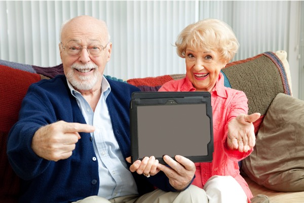 Cheerful elderly couple sitting on a cozy couch at home, smiling and pointing excitedly at a blank tablet screen they are holding together.