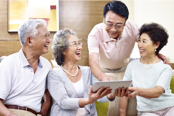 Joyful multi-generational Asian family — two senior couples and their adult son — gathered on a couch, laughing together while looking at a tablet screen in a cozy living room.